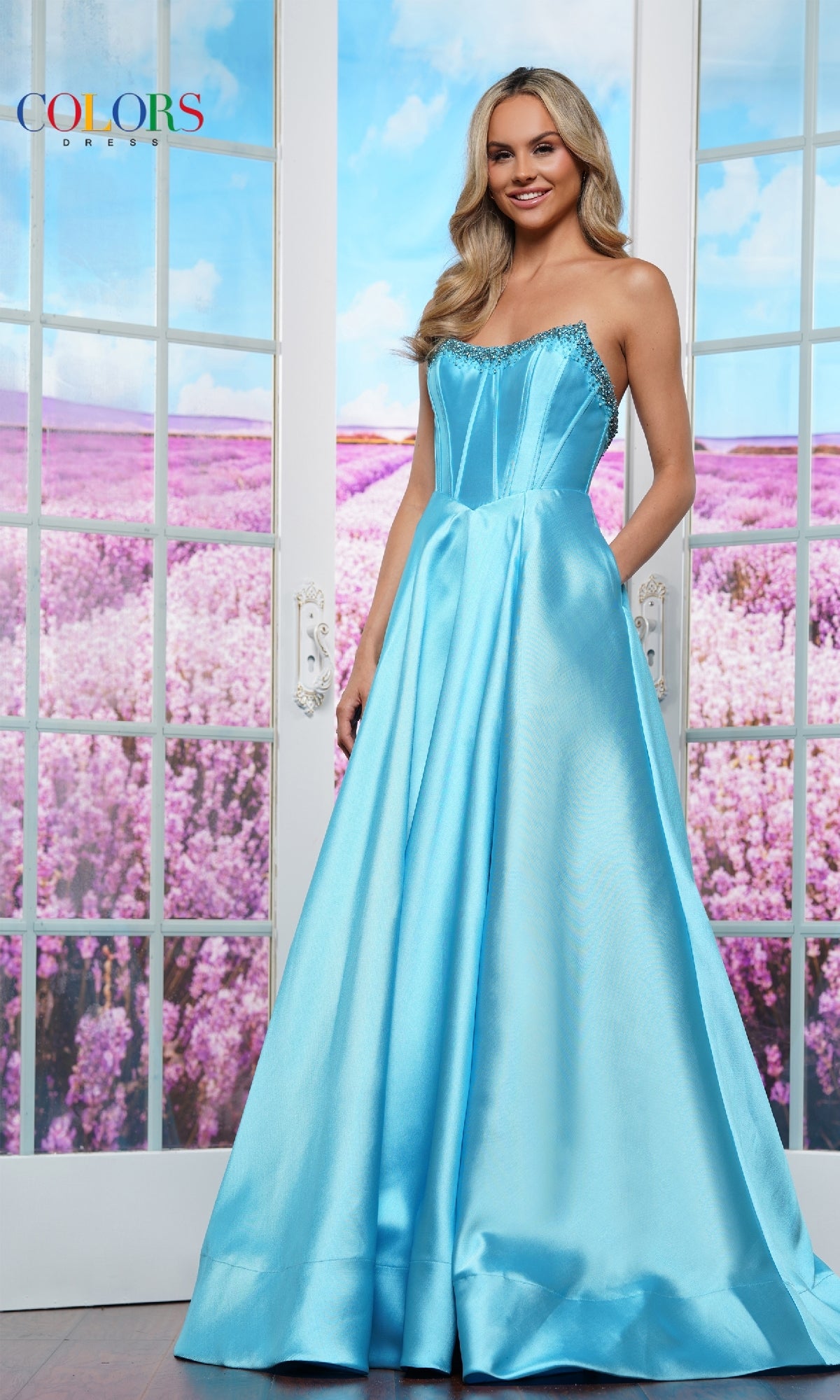 A woman in the Strapless Corset Prom Ball Gown: Colors Dress 3478 stands indoors before glass doors, with a field of purple flowers visible outside.